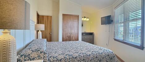 A bedroom with a bed, a dresser, a lamp, a TV, and a window with blinds.
