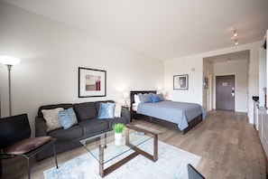 A modern living room with a sofa, a glass coffee table, and a bed with a blue bedspread.