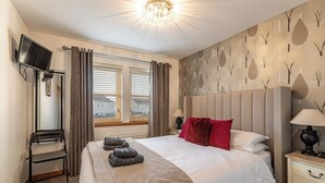 Een net opgemaakt bed met een witte deken en een rood kussen, een klein nachtkastje met een lamp, een stoel met een handdoek, en een muur met een boommotief.