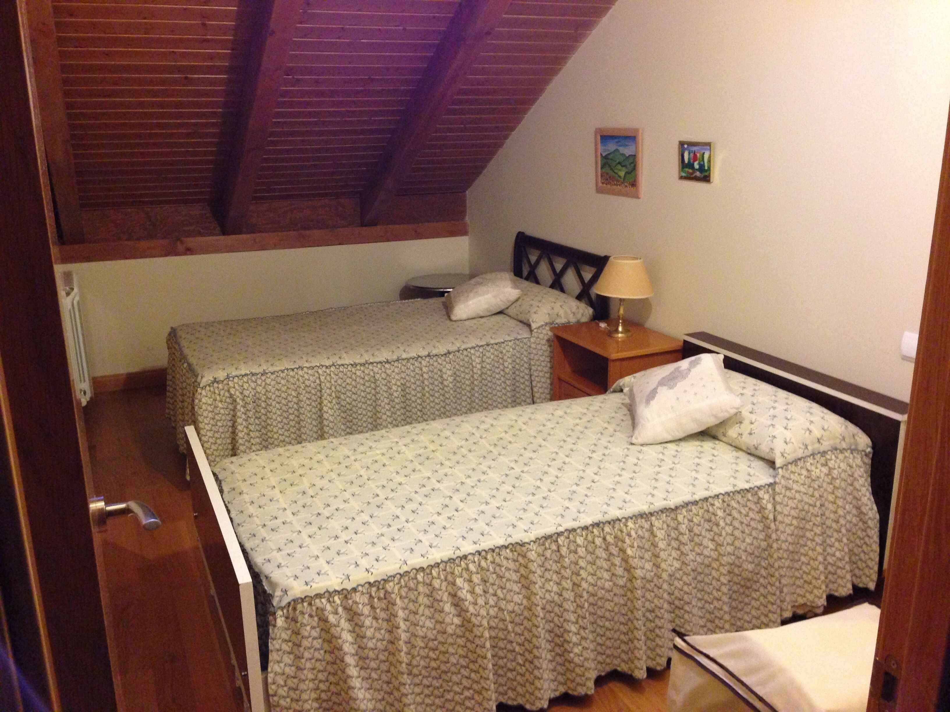 Una habitación pequeña y acogedora con dos camas individuales, techo de madera y una mesita de noche sencilla con una lámpara.