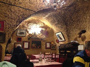 L’intérieur présente un mur en pierre rustique, des tableaux encadrés et un lustre. Les tables sont dressées avec des verres à vin et des assiettes.