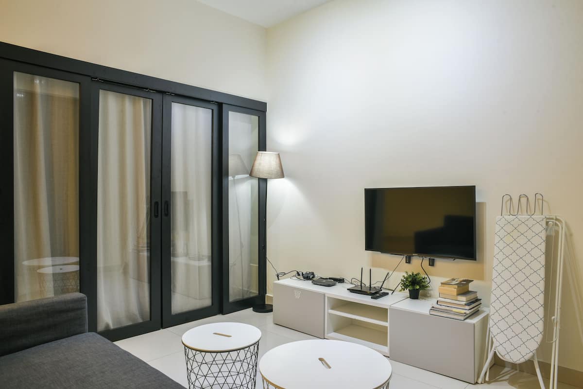 Una sala de estar moderna con un sofá, dos mesitas, un televisor, una lámpara y un mueble blanco con libros y una planta.