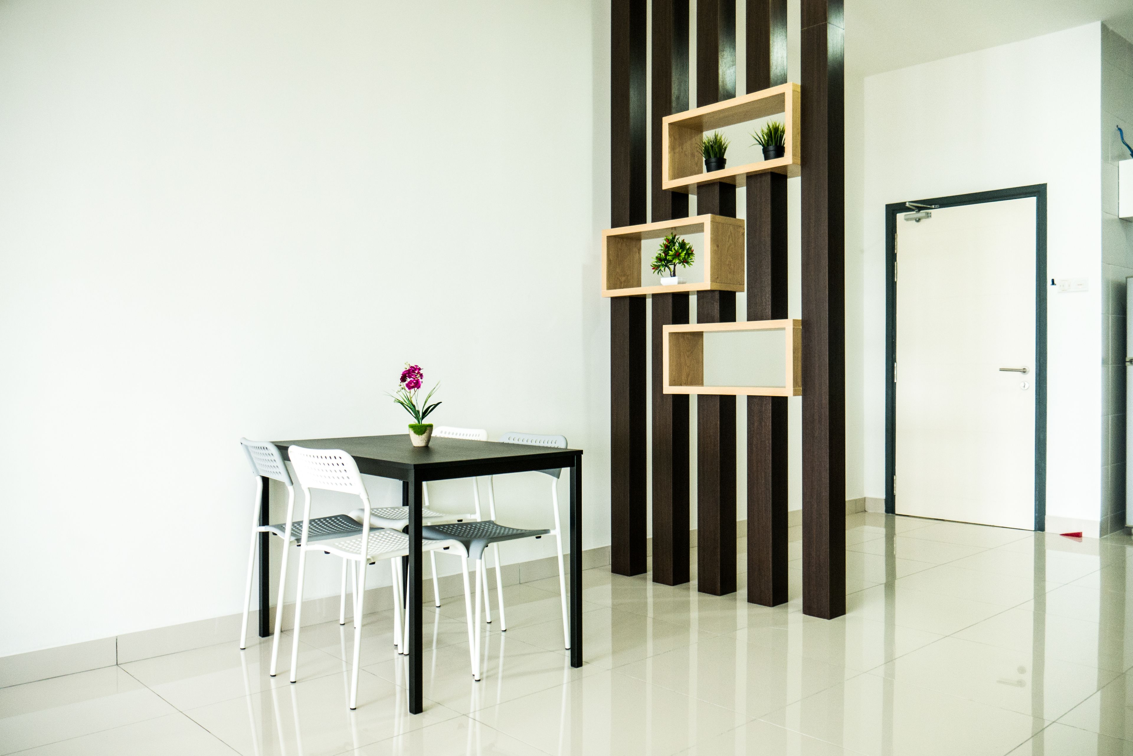 A modern room with a dining table, two white chairs, a potted plant, and a wooden shelving unit with small plants.