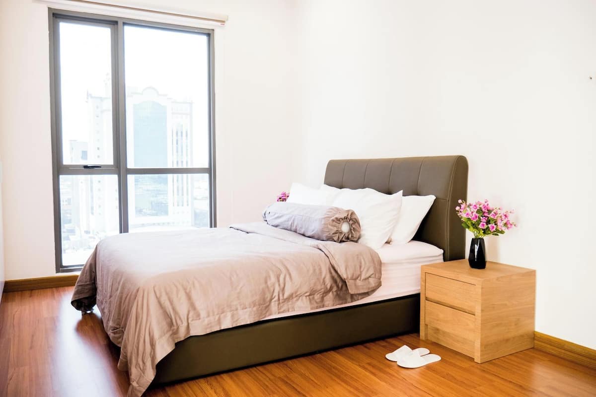 Un dormitorio con cama, mesita de noche, ventana y vistas a edificios.