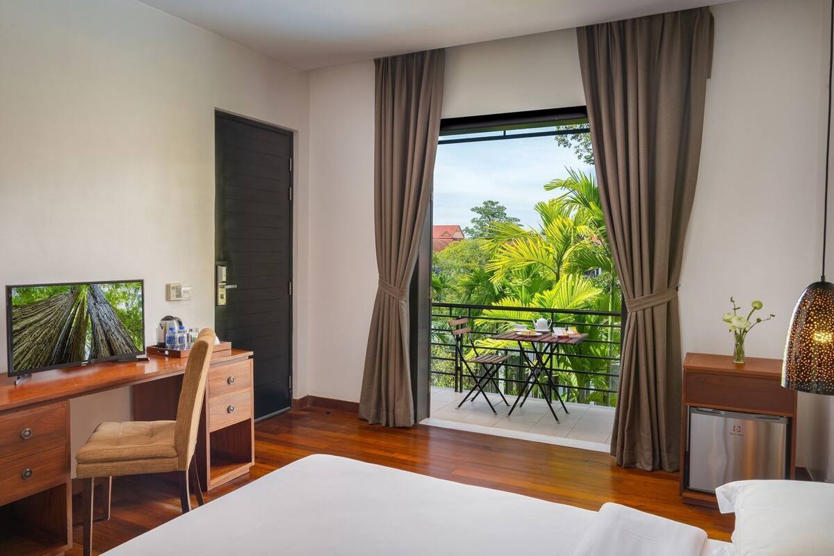 A hotel room with a bed, a desk with a chair, a TV, a balcony with a table and chairs, and a view of greenery.