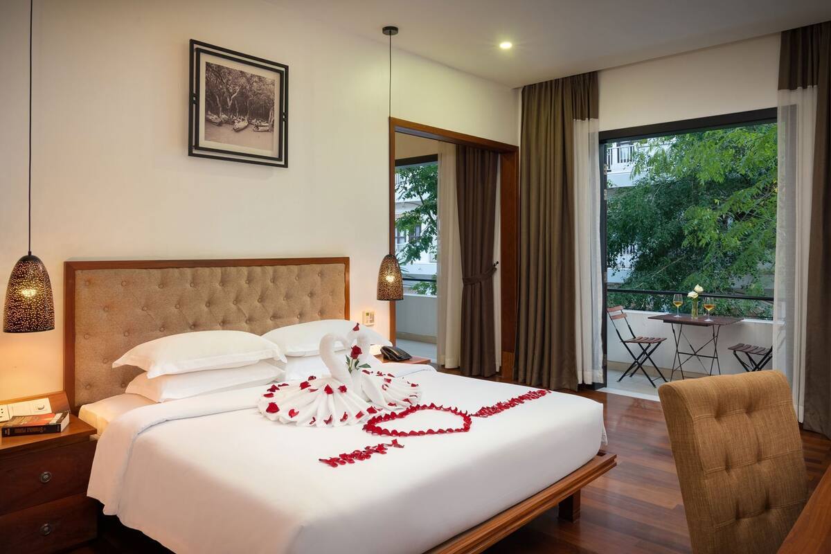 A hotel room with a bed, a dining table with chairs, a view of greenery, and a framed picture on the wall.