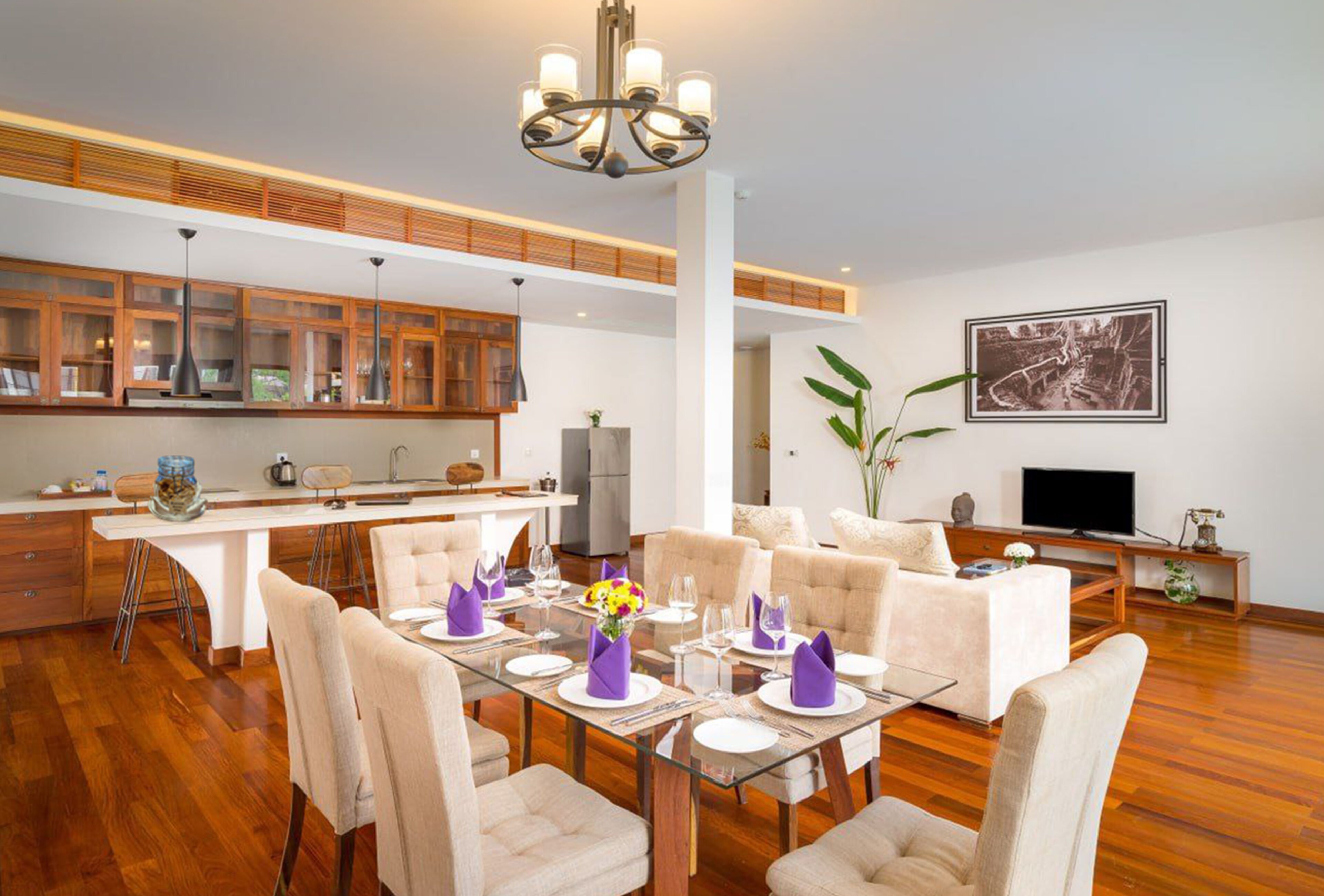 A modern dining area with a glass table set for six, surrounded by beige chairs. A chandelier hangs from the ceiling, and a large framed picture is on the wall.