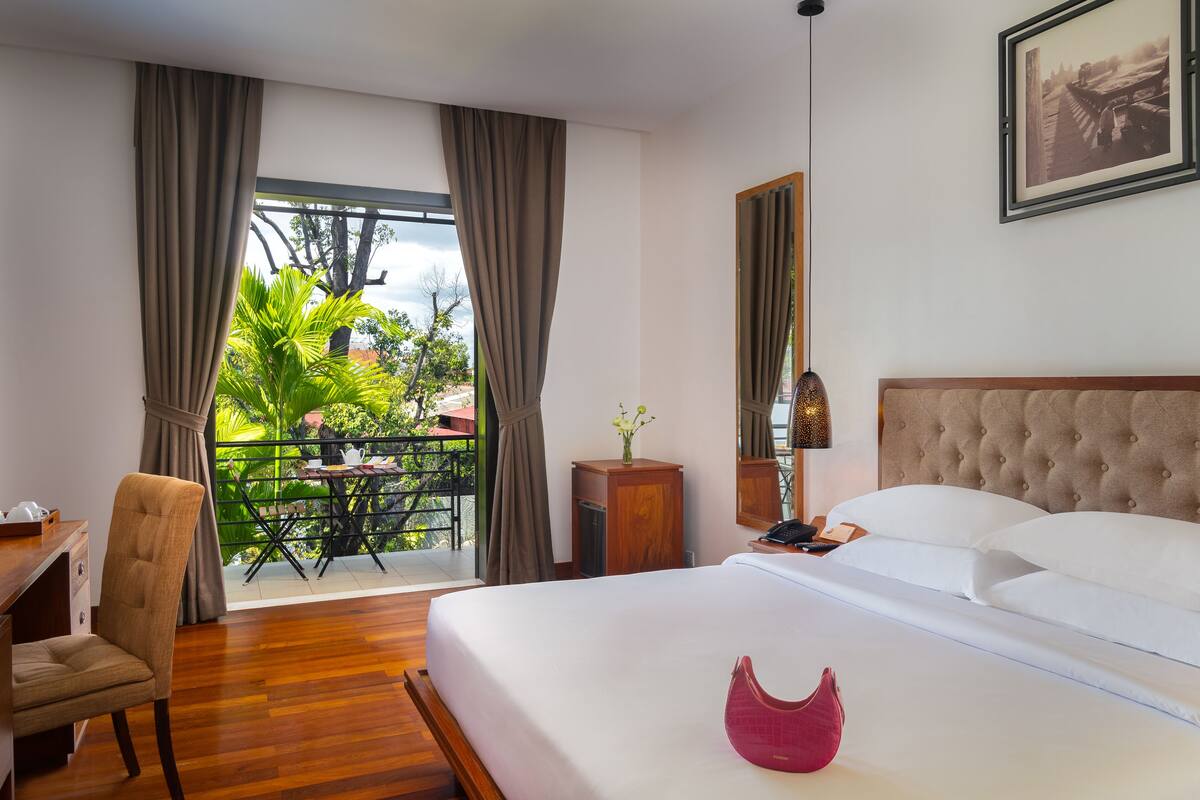 A hotel room with a bed, a desk, a chair, a small table with a vase, and a balcony with plants.