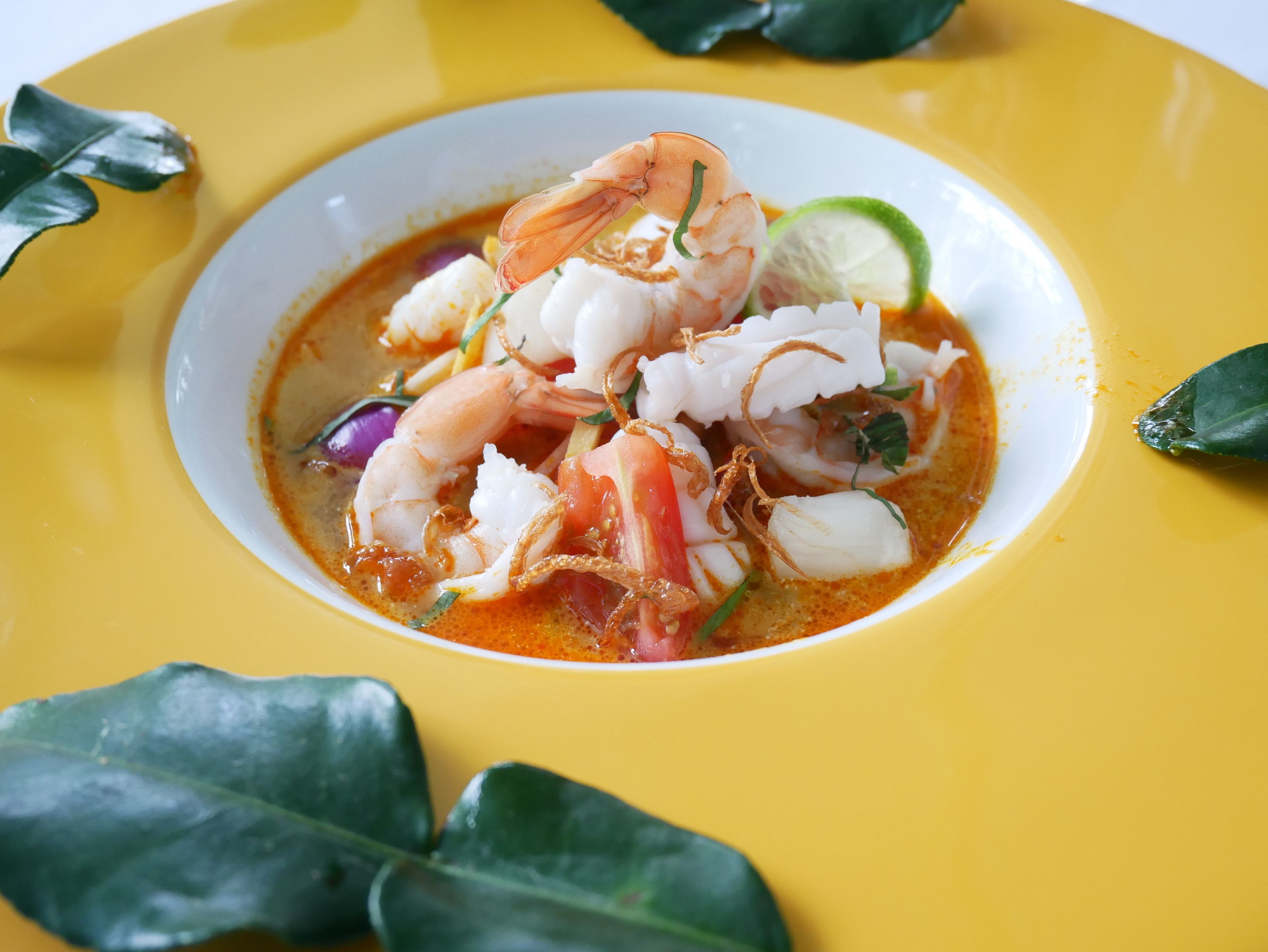 Un tazón de sopa de mariscos con camarones y calamares, adornado con lima y hierbas, servido en un tazón amarillo.