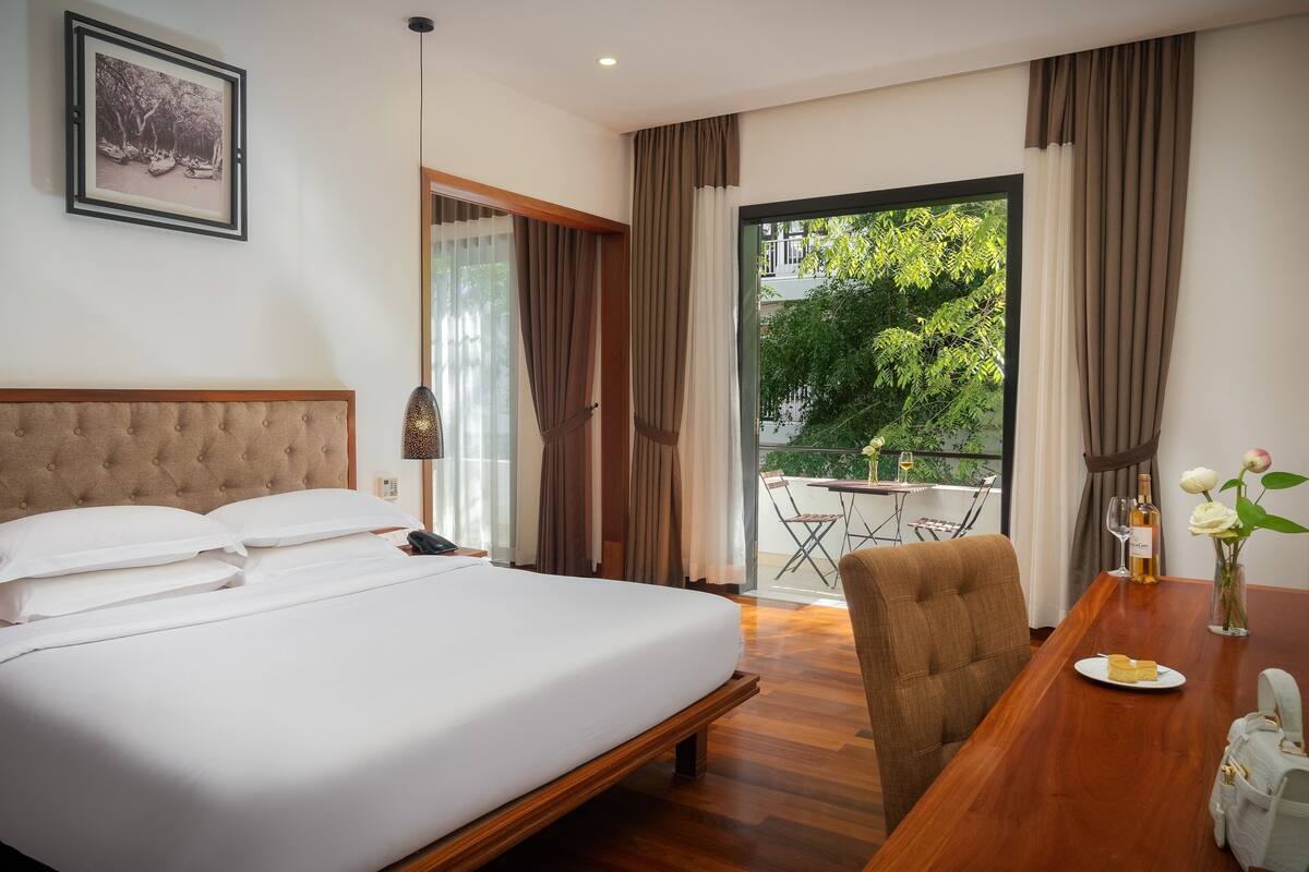 A hotel room with a large bed, a wooden dining table, a chair, a balcony with a table and chairs, and a framed picture on the wall.