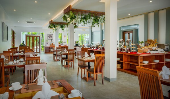 Un comedor amplio con mesas y sillas de madera, un mostrador de buffet con diversas opciones de comida y plantas colgantes.