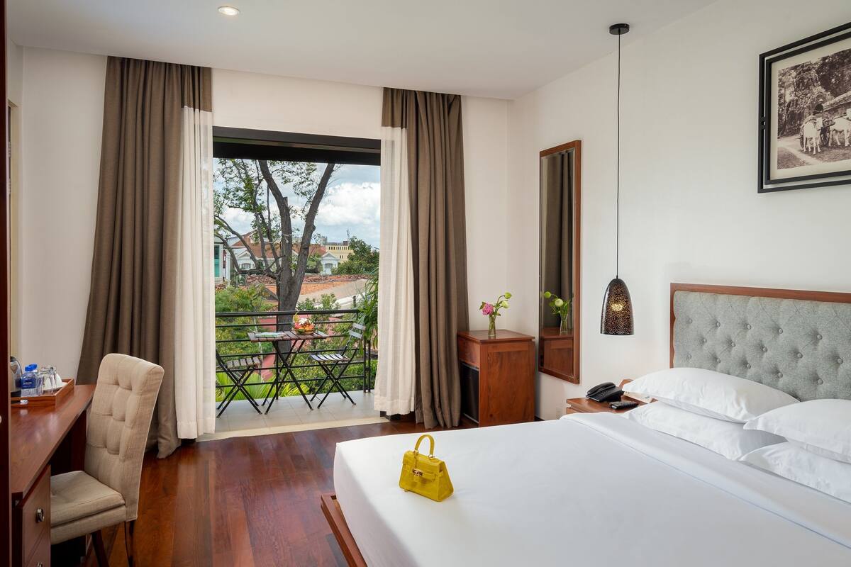 A hotel room with a bed, a desk, a chair, a balcony with a table and chairs, and a framed picture on the wall.