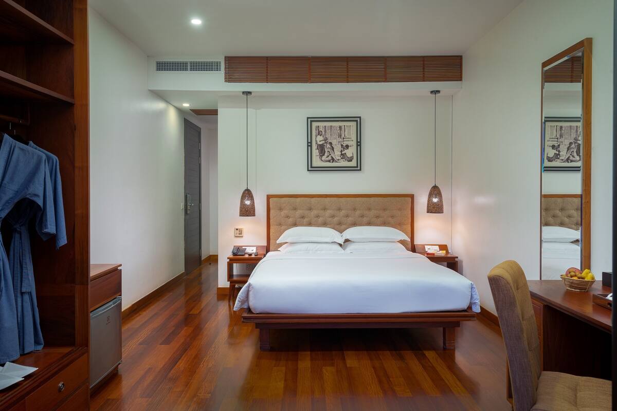 A hotel room with a large bed, wooden flooring, a wardrobe, bedside tables with lamps, and artwork on the walls.