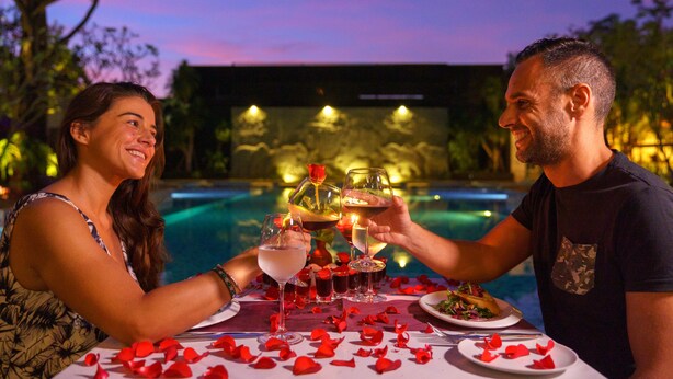 A couple toasting with champagne glasses at a poolside table with rose petals, surrounded by a pool and evening lights.