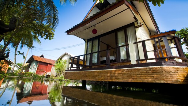 A houseboat with a balcony, situated on a calm water body, surrounded by greenery and other houses. - Lampam Resort (Phatthalung)