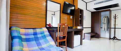 Una habitación con pared de madera, televisor sobre un escritorio, una silla, una mesita, perchero y cama con manta colorida.
