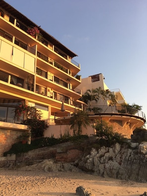 Un hôtel à plusieurs étages doté de balcons, avec une plage en premier plan et des palmiers.