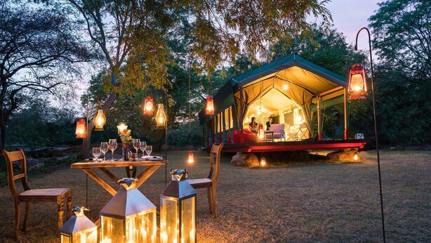 An outdoor dining area with a table set for two, surrounded by lanterns, and a tent with a person inside. - Flameback Eco Lodge (Weerawila)