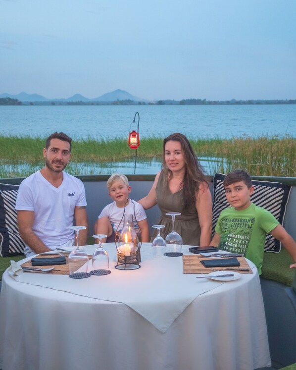 Una familia cenando en una mesa al aire libre con vistas a un lago y montañas al fondo.