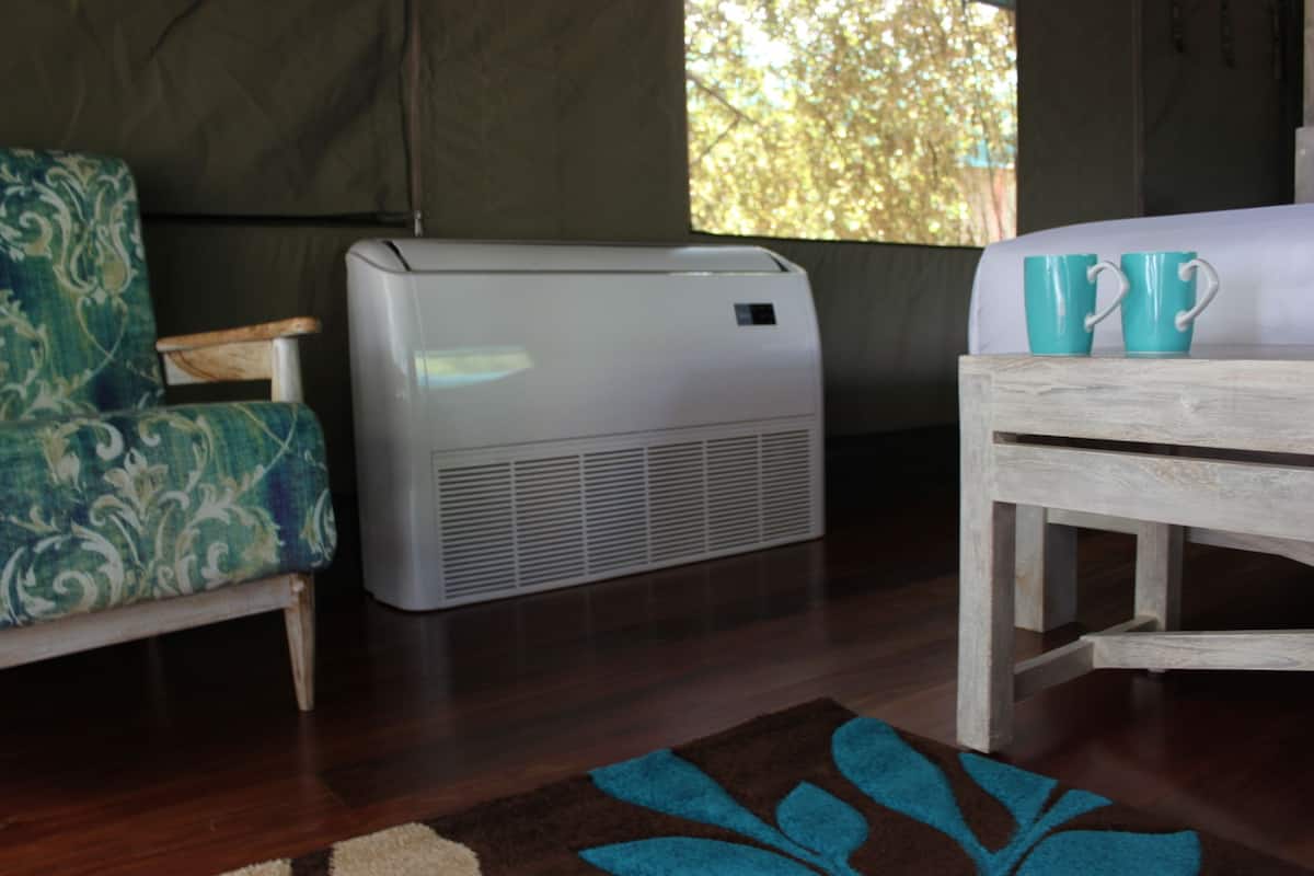 A room with a bed, a table with two teal mugs, a patterned armchair, and a portable air conditioner.