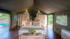 A tented accommodation room with a bed, bedside lamps, a small table with books and a plant, and a view of a swimming pool outside. - Flameback Eco Lodge (Weerawila)