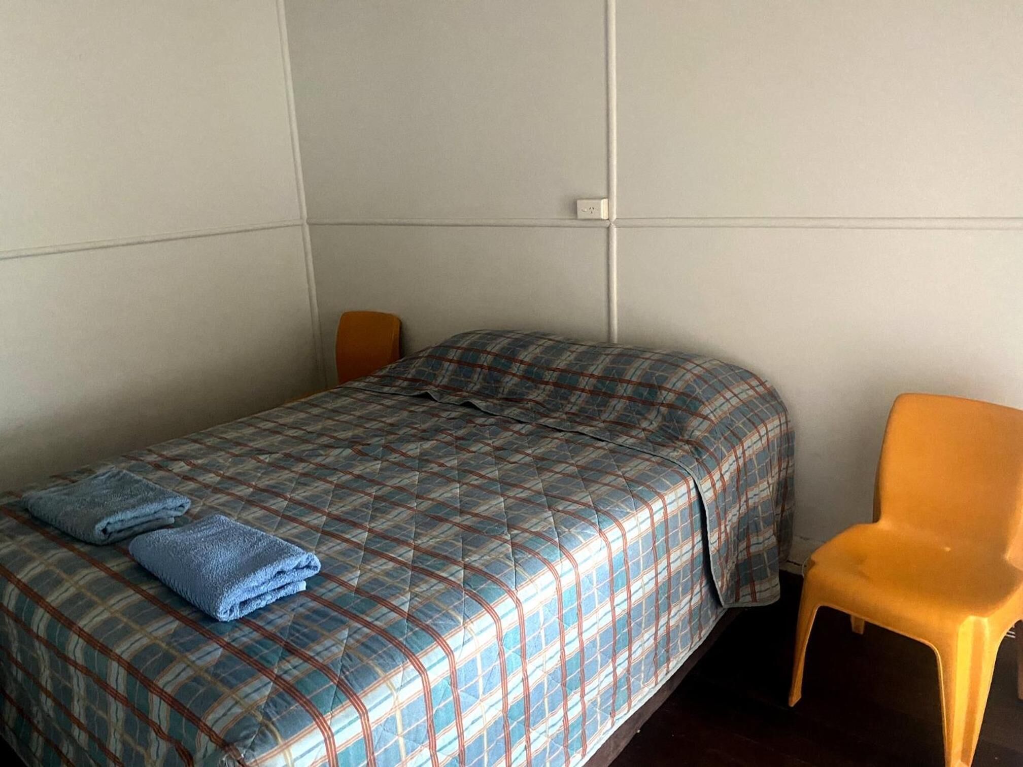 Een enkel bed met een geruite plaid, een opgevouwen blauwe handdoek en een oranje stoel in een kamer met effen muren en een zichtbare stopcontact.'