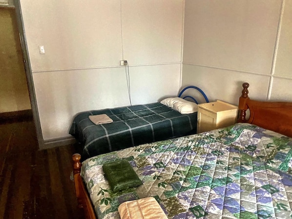 Una habitación pequeña y sencilla con dos camas, una mesita de noche de madera y una pared lisa.