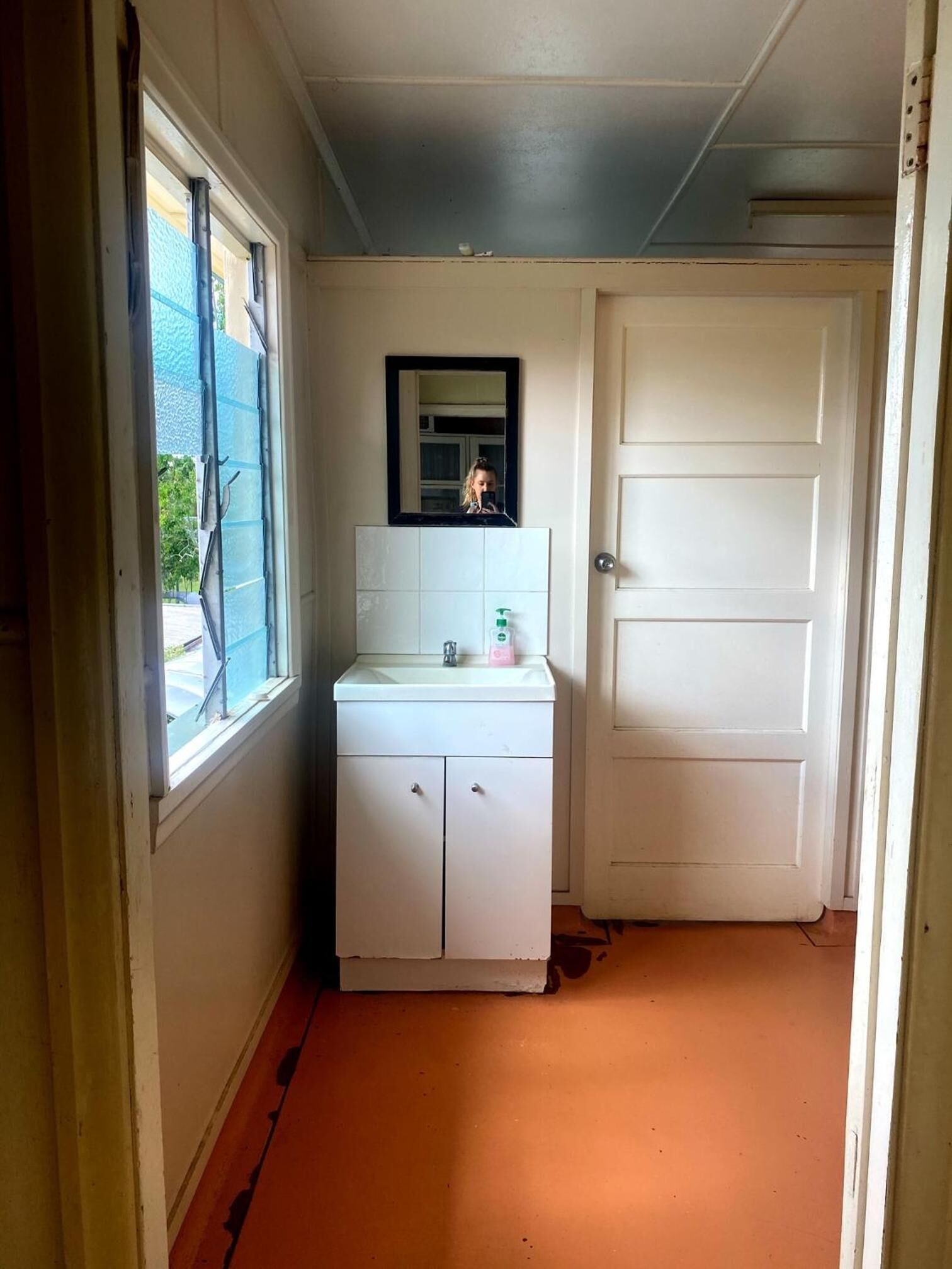 A narrow hallway with a white door, a window with blinds, and a white sink with a mirror above it.