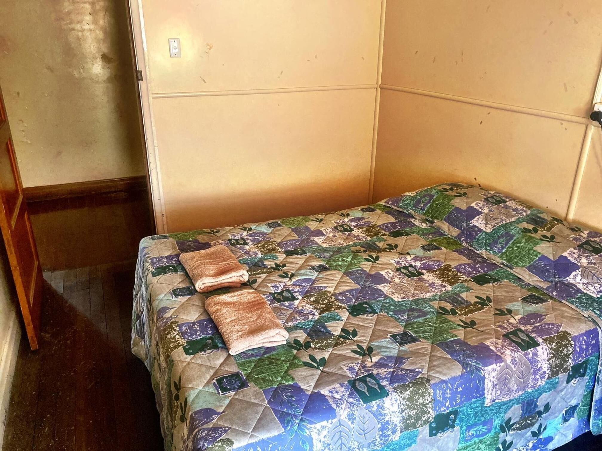A small, single-bed room with a quilted bedspread and two folded towels.