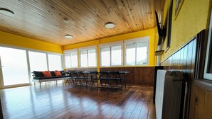 A spacious room with wooden floors, a long table with chairs, a bench, and large windows allowing natural light.