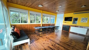 A room with a wooden ceiling, large windows, a dining table with chairs, a sofa, and a radiator.