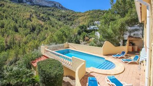 Un espace piscine avec des chaises longues, une vue sur un paysage montagneux et un bâtiment doté d’un toit en tuiles.