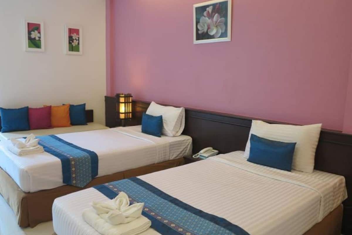 Une chambre d’hôtel avec deux lits, chacun recouvert d’une courtepointe bleue et blanche, avec des oreillers et une serviette pliée. Il y a une table de chevet avec une lampe et un téléphone. Les murs sont peints en rose, et deux tableaux encadrés sont accrochés au mur.
