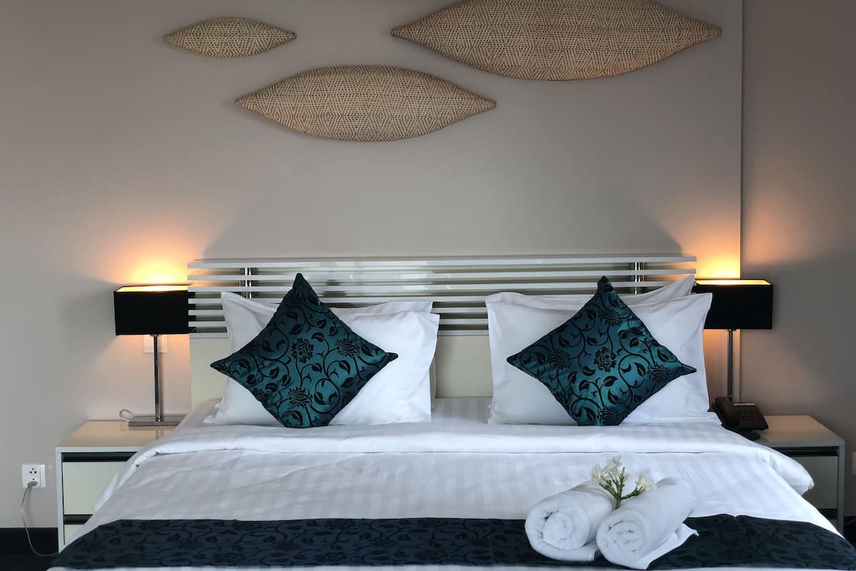A neatly made bed with white linens and two decorative pillows, a bedside table with a lamp, and a wall-mounted decorative piece above the bed.