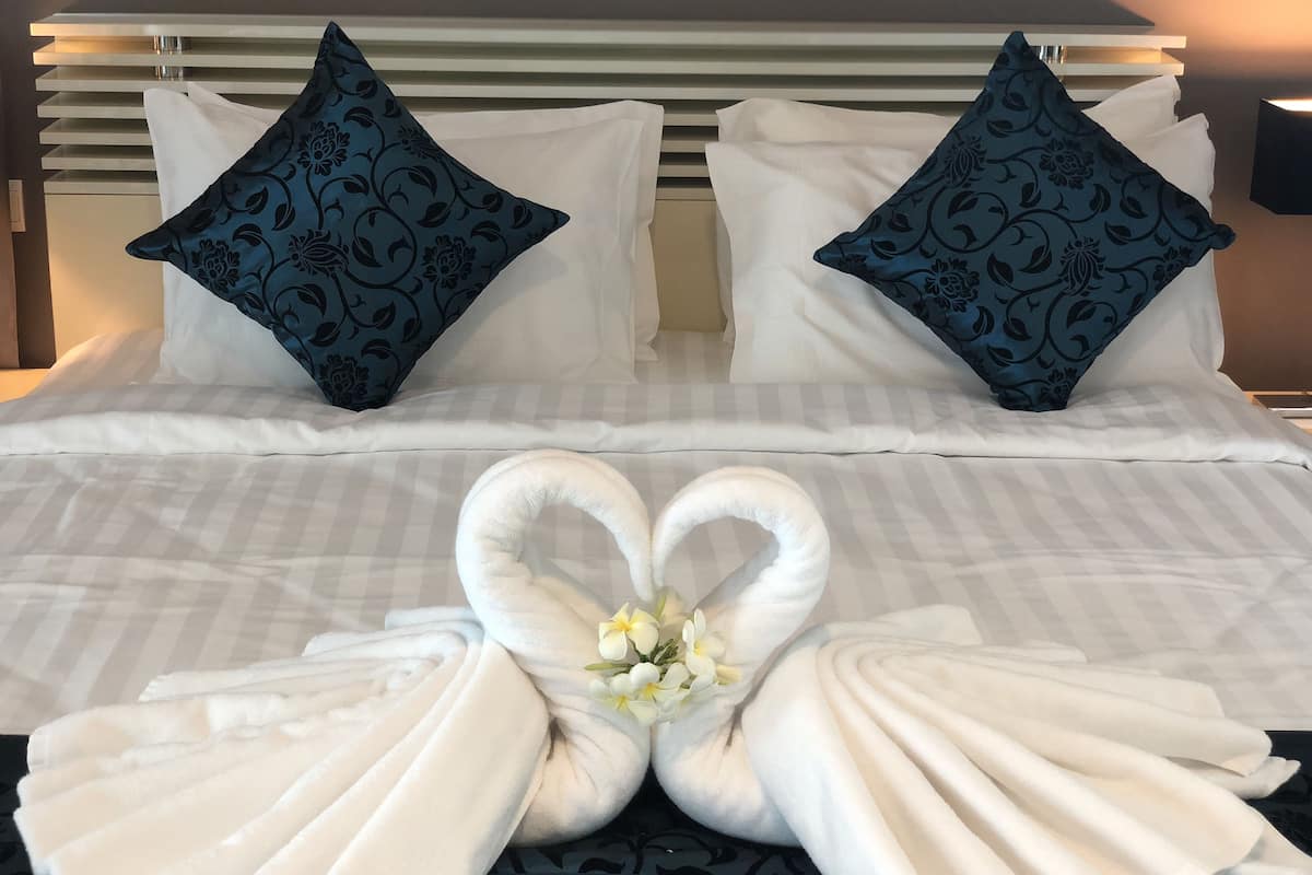 A neatly made bed with white linens, two decorative pillows, and a towel arranged in a heart shape with flowers.