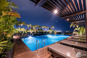 An outdoor swimming pool with a wooden deck, palm trees, and a view of the city at night.