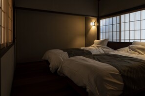 A dimly lit room with a bed, a window with blinds, and a wall-mounted light fixture.