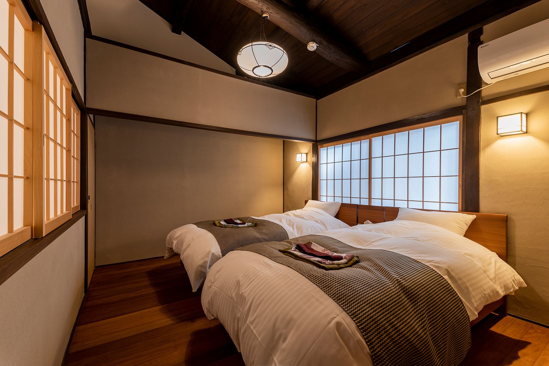 A traditional Japanese-style bedroom with a wooden ceiling, a large window with a grid pattern, and a bed with white bedding and a patterned blanket.
