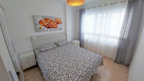 A bedroom with a bed, a nightstand, a painting, a window with curtains, and a hanging light fixture. - Apartamento Sarah 2A, 2min to the beach and promenade, central and quiet. Free WIFI (Morro Jable)