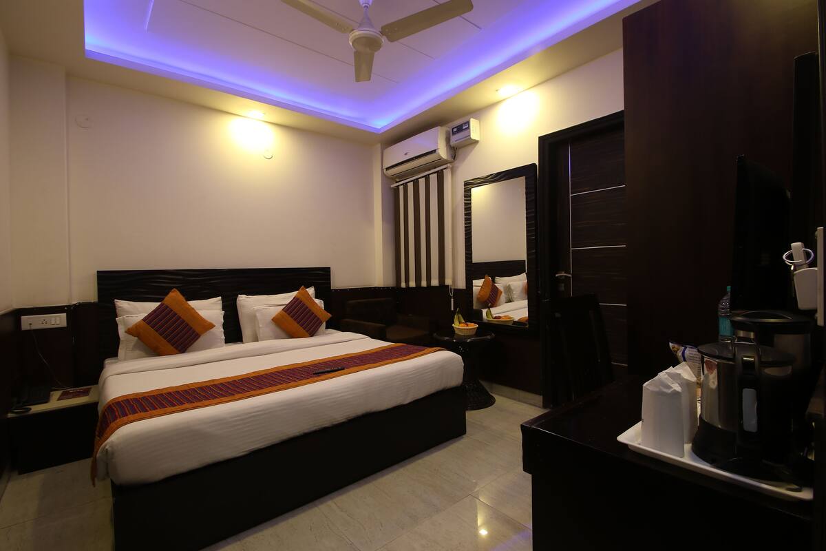 A hotel room with a large bed, a bedside table with a lamp, a coffee maker, and an air conditioning unit.