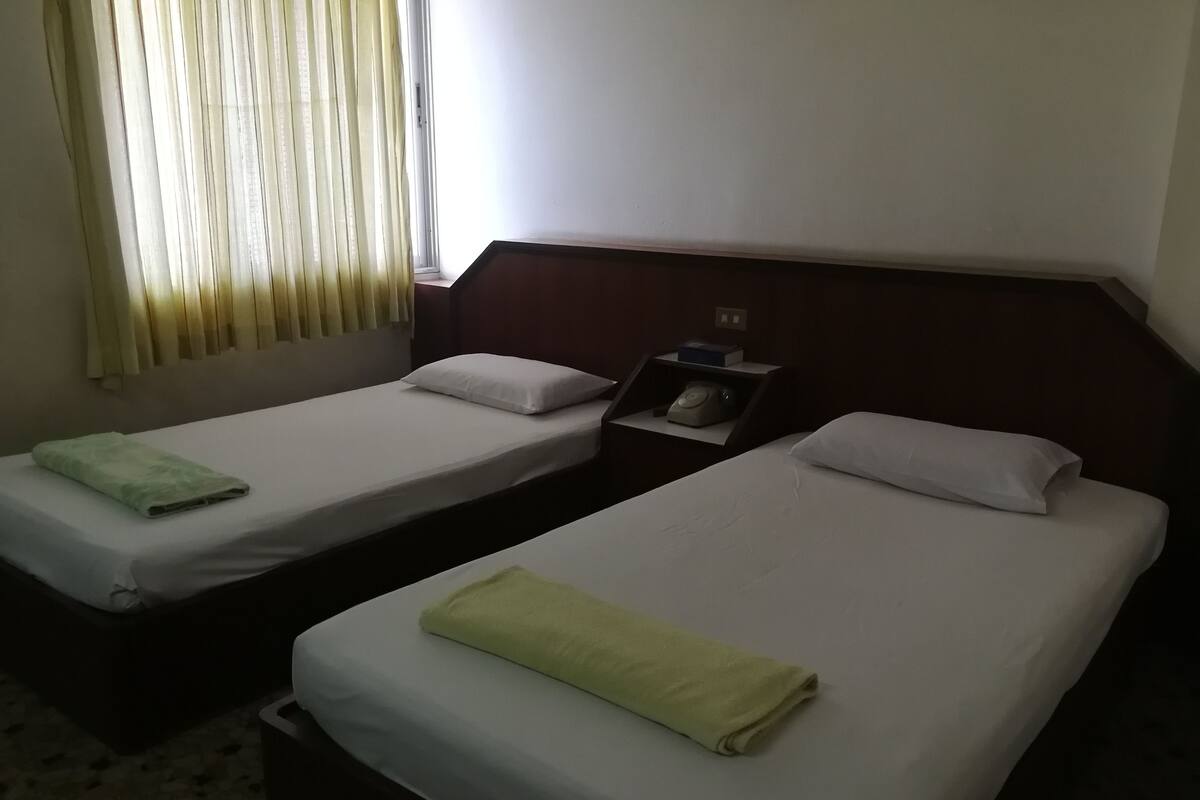 Habitación de hotel con dos camas individuales, una mesita de noche con teléfono y una ventana con cortinas.