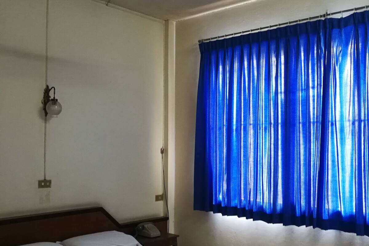 Habitación de hotel con una cama individual, ventilador de techo, mesita de noche con lámpara y ventana con cortinas azules.