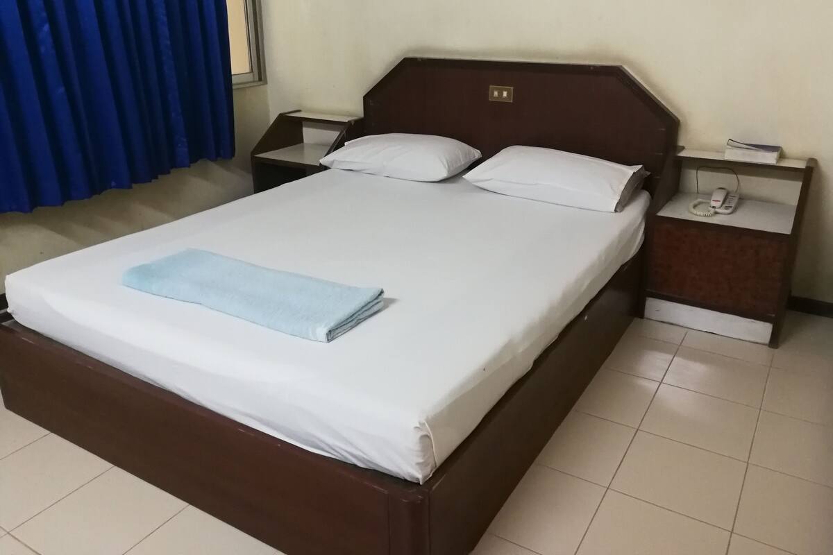 Una cama bien tendida con sábanas blancas y una toalla azul, un cabecero de madera, dos mesitas de noche con lámparas y un teléfono.