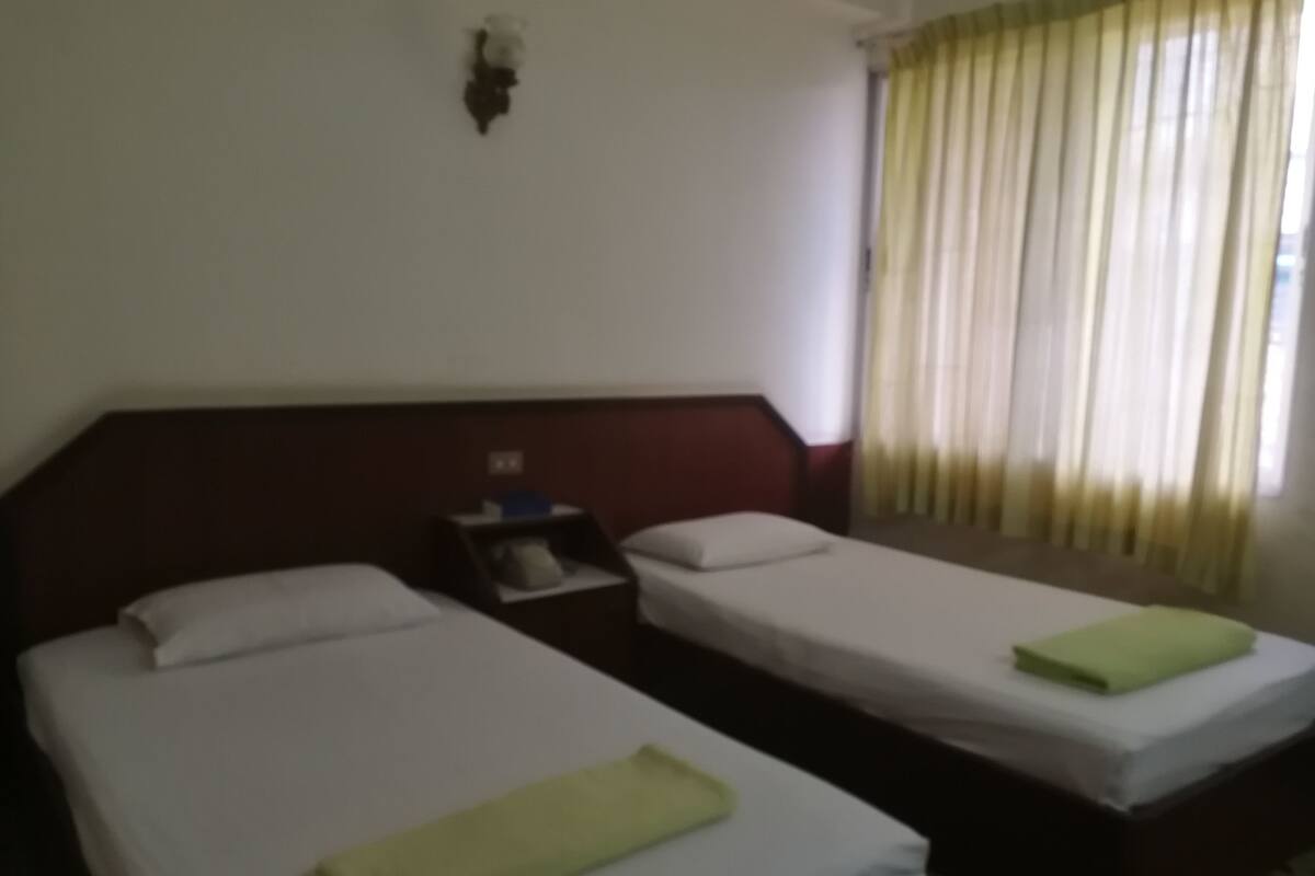Habitación de hotel con dos camas individuales, una mesita de noche y una ventana con cortinas.