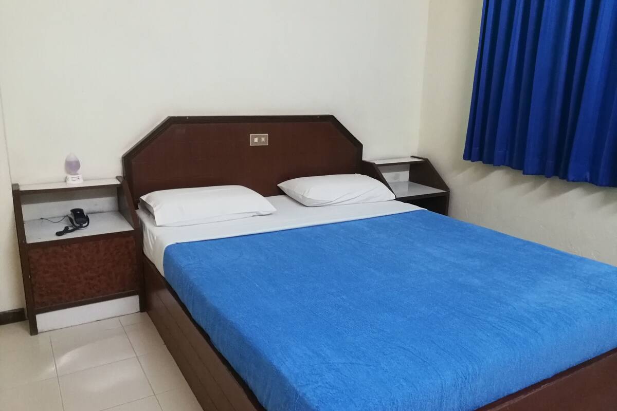 Una cama bien hecha con una colcha azul, dos almohadas blancas y un cabecero de madera. Se ve una mesita de noche con una lámpara y una pequeña mesa junto a la cama.