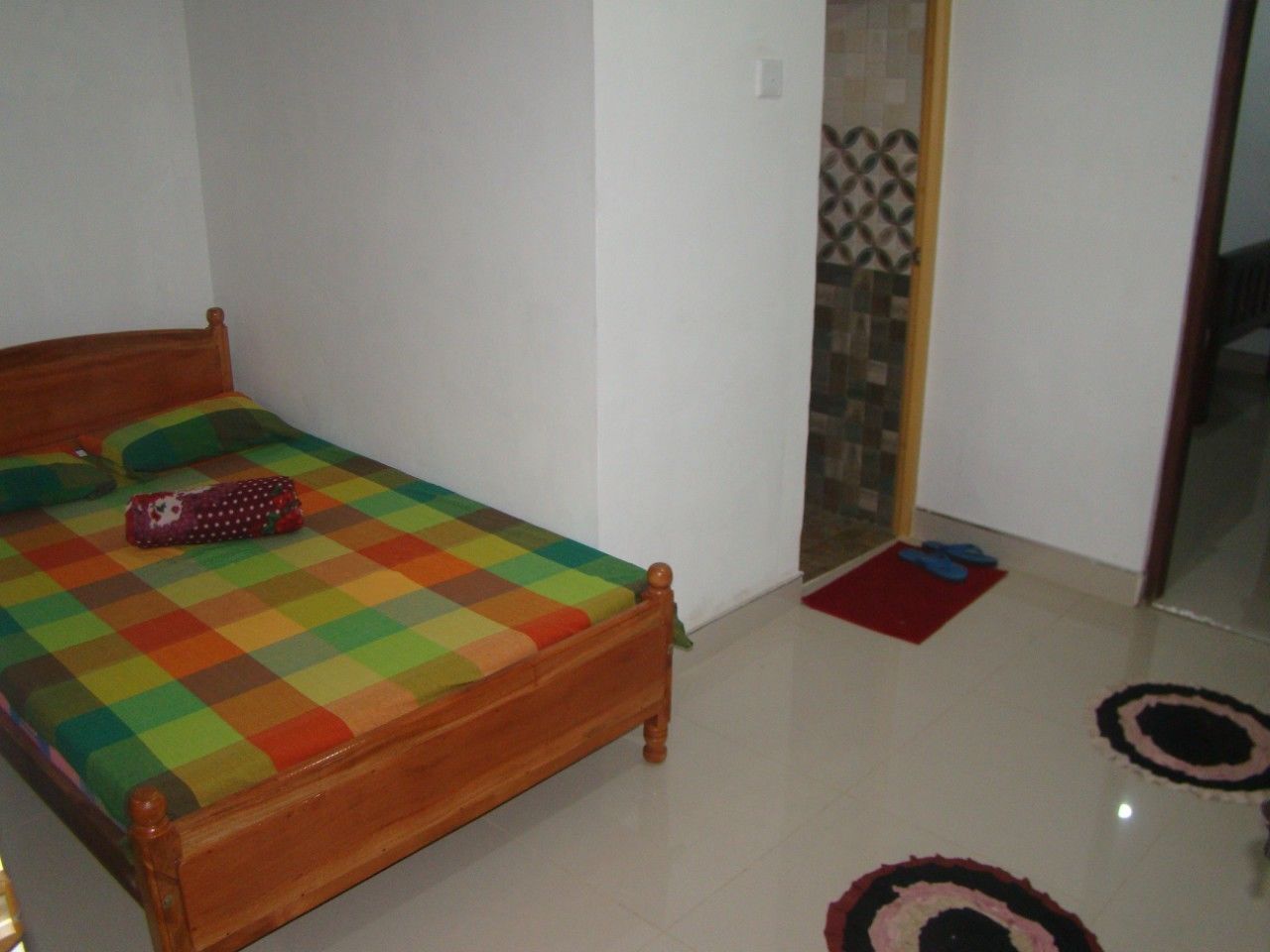 A bedroom with a wooden bed, a colorful checkered bedspread, and a small red rug on the floor.