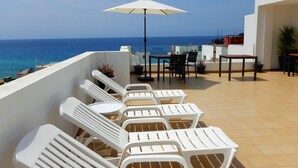 Eine Terrasse mit weißen Kunststoffliegen und einem Tisch, mit Blick auf das Meer.
