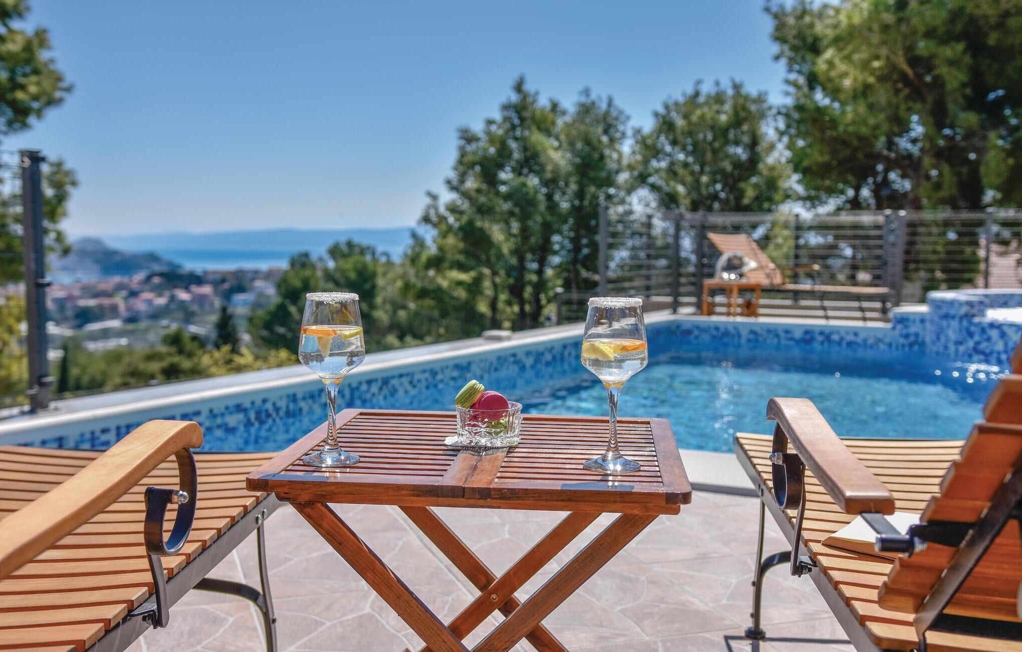 A poolside table with two glasses of a clear beverage, a colorful macaroon, and a view of a pool and landscape.