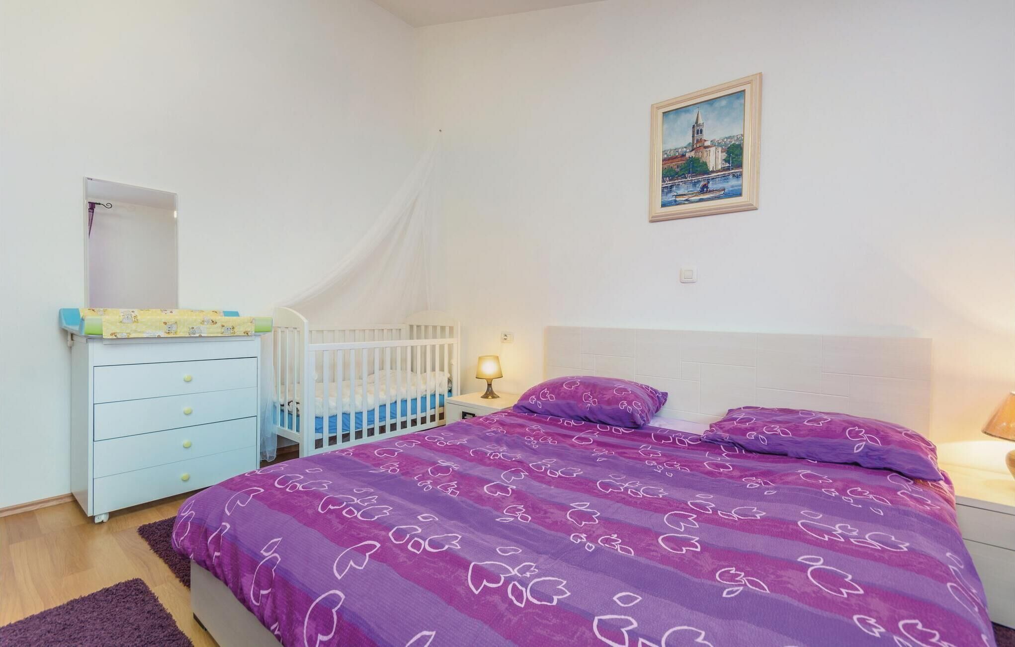 Une chambre avec un berceau, un lit recouvert d’une courtepointe à motifs floraux violets, une table de chevet avec une lampe et un tableau représentant un paysage urbain accroché au mur.
