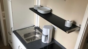 A compact kitchen with white cabinets, a black countertop, a built-in stove, a coffee maker, and shelves holding plates and cups.